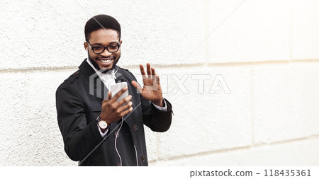 Black businessman have video call on the way to office, waving at phone screen, standing over white wall outdoors, copy space Black businessman have video call on the way to office, waving at phone screen, standing over white wall outdoors, copy space 118435361