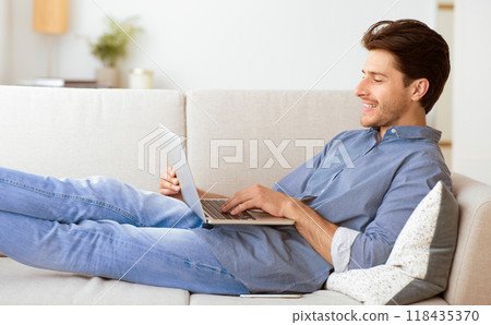 A man is lying on a white couch, using a laptop, while smiling. He is dressed in blue jeans and a blue button-down shirt. 118435370