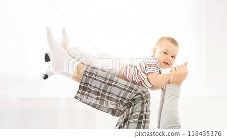 An image of a father holding his baby son in the air. The father is wearing plaid pajama pants and the baby is wearing a striped shirt. Both are smiling. 118435376