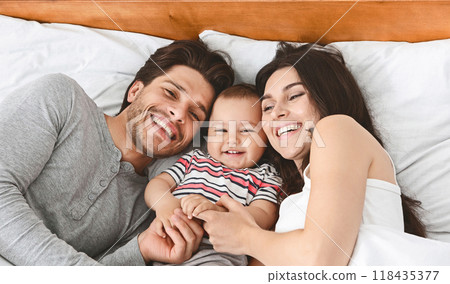 A happy family of three lies in bed together, smiling and laughing. The parents are both looking at their baby, who is lying between them. The baby is also smiling and laughing. 118435377