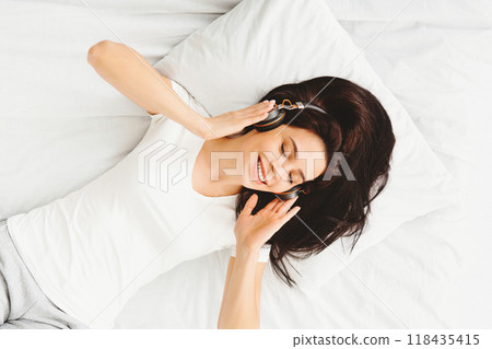 Personal relax. Young millennial woman listening to music, lying in bed in wireless headphones, top view 118435415