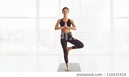 A woman maintains a steady tree pose, a yoga asana, balancing on one leg with the other foot placed against her inner thigh. Her hands are pressed together in a prayer position 118435419