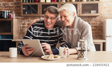 A senior couple is sitting together at a table in their kitchen. The man is looking at a tablet while the woman is smiling and looking at him. They appear to be sharing a moment of joy and laughter A senior couple is sitting together at a table in their kitchen. The man is looking at a tablet while the woman is smiling and looking at him. They appear to be sharing a moment of joy and laughter 118435421