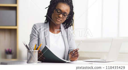 Corporate black businesswoman reviewing documents at her desk in a modern office environment, working diligently. 118435429