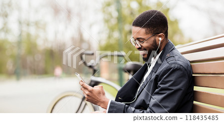 An African American businessman is using his phone while sitting on a park bench with his bicycle nearby. The mood is relaxed and professional, set in a green, serene park. An African American businessman is using his phone while sitting on a park bench with his bicycle nearby. The mood is relaxed and professional, set in a green, serene park. 118435485
