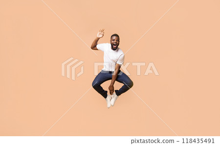 A Black man is jumping in mid-air with his arms raised and an ecstatic expression on his face. He is wearing a white t-shirt and blue jeans. The background is a peach color. 118435491