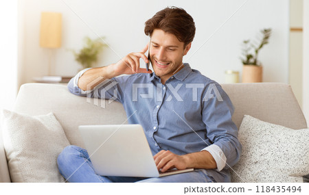 A man sits on a sofa, using a laptop and talking on his phone. He appears to be working from home. A man sits on a sofa, using a laptop and talking on his phone. He appears to be working from home. 118435494