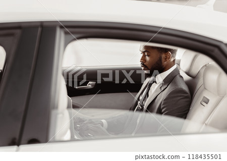 Black man in a suit is sitting in the back seat of a white car. He is looking down and appears to be thinking or working on something. The car is parked in a bright location, likely outdoors. Black man in a suit is sitting in the back seat of a white car. He is looking down and appears to be thinking or working on something. The car is parked in a bright location, likely outdoors. 118435501