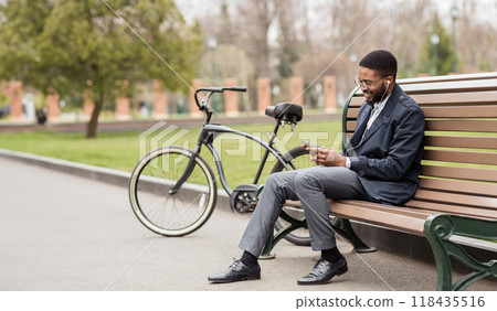 Black businessman is talking on his phone while sitting on a bench in the park, with a bicycle nearby. The atmosphere is relaxed and efficient, set in a modern outdoor environment. 118435516
