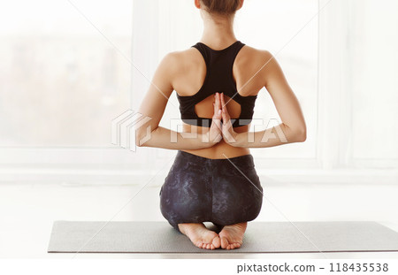 A young woman is engaged in a yoga session, with her back facing the camera, sitting on a yoga mat with her feet tucked under and her hands are pressed together in a prayer position behind her back A young woman is engaged in a yoga session, with her back facing the camera, sitting on a yoga mat with her feet tucked under and her hands are pressed together in a prayer position behind her back 118435538