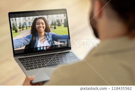 African American woman is on a video call with someone on a laptop computer. She is smiling and waving, and appears to be in a park setting. She is likely calling a friend African American woman is on a video call with someone on a laptop computer. She is smiling and waving, and appears to be in a park setting. She is likely calling a friend 118435562