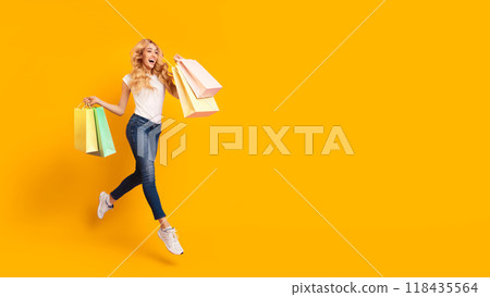 A woman in jeans and a white t-shirt jumps in the air with excitement, holding shopping bags in her hands against a yellow background. She is smiling widely, with her blond hair flowing behind her. 118435564