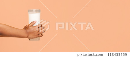 Baby hand is holding a glass of milk against a peach-colored background. The glass is filled with white milk, and the hand is shown from the wrist to the fingertips, panorama with copy space Baby hand is holding a glass of milk against a peach-colored background. The glass is filled with white milk, and the hand is shown from the wrist to the fingertips, panorama with copy space 118435569