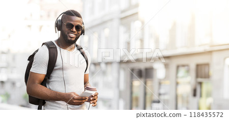 A man with headphones and a backpack walks down the street, looking at his phone. 118435572