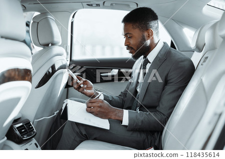 Black man in a suit sits in the backseat of a car and works on his smartphone while writing notes in a notebook. He appears to be focused and engaged in his work 118435614