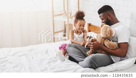 African American father and his daughter are sitting on a bed in a bedroom. They are both smiling and playing with stuffed animals, copy space African American father and his daughter are sitting on a bed in a bedroom. They are both smiling and playing with stuffed animals, copy space 118435635