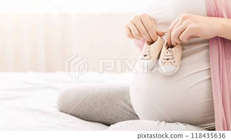 Happy pregnancy. Mother holding small baby shoes on pregnant belly, empty space Happy pregnancy. Mother holding small baby shoes on pregnant belly, empty space 118435636