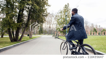 Black businessman is cycling near a green public park. The mood is dynamic and calm, showing a seamless transition between urban work life and natural relaxation. 118435647