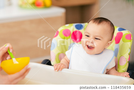 A mom is feeding her baby boy in a high chair in the kitchen. The mood is warm and affectionate, with a cozy kitchen environment enhancing the loving scene. 118435659
