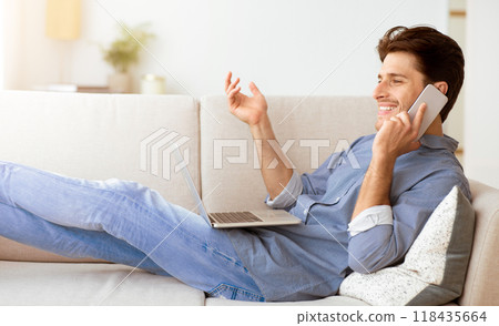 A man sits on a couch with a laptop in his lap and talks on a phone with a smile on his face. 118435664