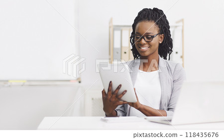 Positive young black lady wearing formal outfit and eyeglasses manager sitting at desk with laptop, using digital tablet at office, copy space Positive young black lady wearing formal outfit and eyeglasses manager sitting at desk with laptop, using digital tablet at office, copy space 118435676