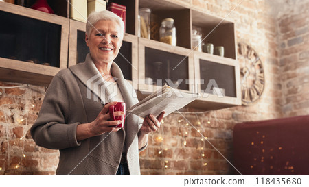A woman with gray hair is standing in her kitchen, smiling and holding a newspaper in one hand and a red mug of coffee in the other, copy space 118435680