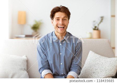 A smiling man sits on a white couch in a living room, wearing a blue button-up shirt. 118435685