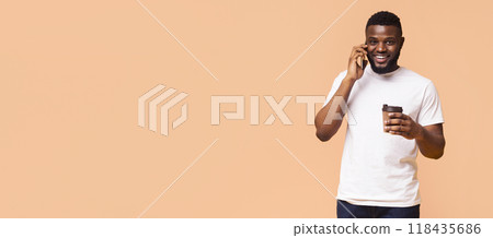 A happy young black man with a beard is smiling while holding a coffee cup in one hand and talking on his phone with the other. He is wearing a white t-shirt, standing against a pale background. 118435686