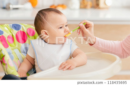 A woman is sitting and feeding a baby with a spoon. The baby is sitting on a high chair while eagerly eating the food. The woman gently guides the spoon into the baby mouth. 118435695