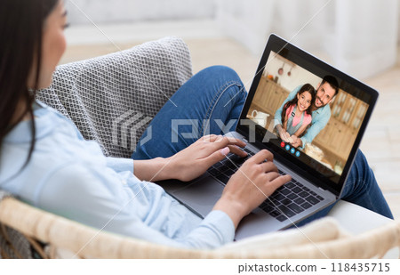Lady sits on a couch in her living room, using a laptop to video chat with her family. She appears to be smiling and happy, enjoying the connection with her loved ones. 118435715