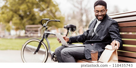 A professional black man uses his laptop on a park bench with a bicycle nearby. The atmosphere is serene and efficient, capturing the essence of outdoor productivity. 118435743