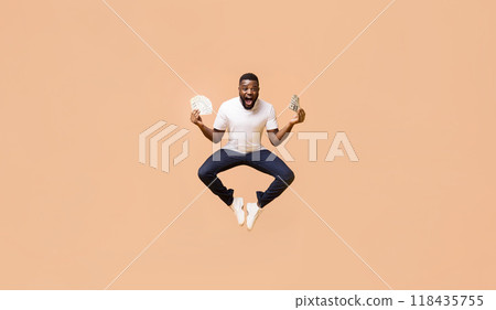 African American young man in a white t-shirt and dark jeans is jumping in the air, with his arms outstretched, holding stacks of money in each hand. The background is a soft peach color. 118435755