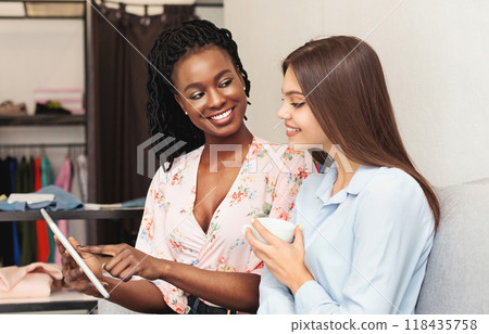 Two multiracial women are standing side by side, both focused on a tablet they are holding. They appear engaged in whatever is on the screen, possibly discussing or sharing information. 118435758