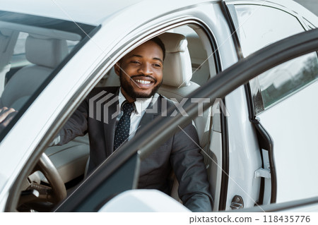 A young African American businessman, dressed in a suit and tie, is smiling as he enters a white sedan car. The car door is open, and the mans hand is on the steering wheel 118435776