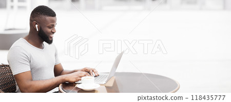 A man wearing earbuds sits at a table in a cafe, smiling while working on his laptop. 118435777