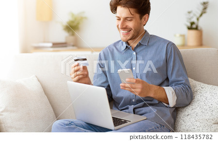A man on a couch smiles while looking at his phone holding a credit card, with a laptop in front, implying hes making an online payment. 118435782