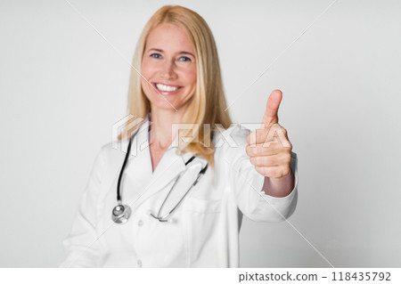 A blonde woman doctor wearing a white coat and a stethoscope gives a thumbs up gesture while smiling. She is photographed against a white background. A blonde woman doctor wearing a white coat and a stethoscope gives a thumbs up gesture while smiling. She is photographed against a white background. 118435792