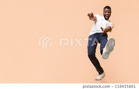 A young Black man in a white t-shirt and blue jeans is dancing in front of a peach-colored background. He has a wide smile on his face and is lifting his right leg, copy space A young Black man in a white t-shirt and blue jeans is dancing in front of a peach-colored background. He has a wide smile on his face and is lifting his right leg, copy space 118435801