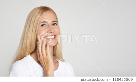 A young blonde woman with a bright smile poses in front of a plain white background. She is wearing a white t-shirt and has her hand gently resting on her cheek, copy space A young blonde woman with a bright smile poses in front of a plain white background. She is wearing a white t-shirt and has her hand gently resting on her cheek, copy space 118435809