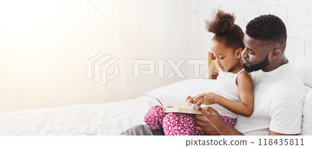 A little Black girl sits on her fathers lap as they read a book together in bed. The father is smiling and looking at his daughter, who is pointing at the book. The scene is warm and loving 118435811