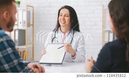 Woman doctor wearing a white coat is sitting at a table and discussing medical information with a couple. She is holding a clipboard and pointing at it with a pen. 118435812