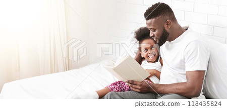 Black father and daughter are sitting on a bed in a bedroom, reading fairytale together. They seem to be enjoying spending time together, panorama with copy space Black father and daughter are sitting on a bed in a bedroom, reading fairytale together. They seem to be enjoying spending time together, panorama with copy space 118435824