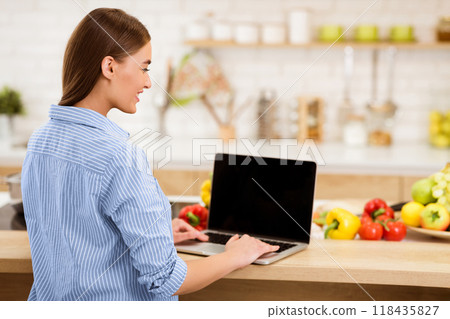 A woman wearing a blue striped shirt uses a laptop while standing at a kitchen counter. Fresh produce, like peppers and tomatoes, are in the foreground. 118435827