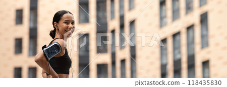 Fit Young Woman In Stylish Sportswear Resting Outdoors After Training, Sporty Athletic Lady With Earphones And Armband Phone Holder Standing On Urban City Street And Smiling At Camera, Copy Space 118435830