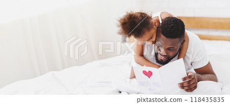 African American little daughter is giving her father a card that says Happy Fathers Day while they lay in bed together. The father is smiling and looking at the card, copy space 118435835
