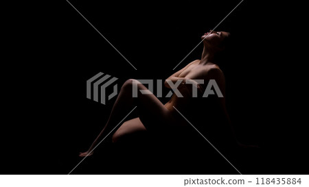 This image shows a woman reclining in a dark room. She is sitting with her legs extended and her head tilted back, seemingly in a state of contemplation or relaxation. 118435884