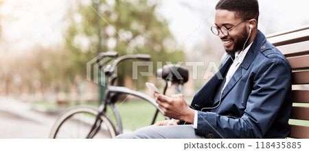 An African American man is using his phone while sitting on a bench in the park with his bicycle close by. The setting is outdoors, highlighting a peaceful yet professional atmosphere. 118435885