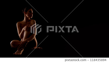 A woman sits alone in a dark room, her arms crossed and her legs crossed. The only light is from the side, illuminating her profile and creating a sense of mystery, copy space 118435894