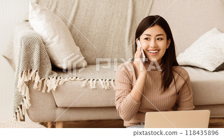 Young Freelancer Talking on Phone and Using Laptop, Sitting on Carpet at Home 118435896