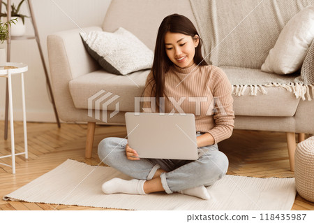 A woman sits on a rug in a living room, using a laptop computer. She is smiling and appears to be focused on her work. A woman sits on a rug in a living room, using a laptop computer. She is smiling and appears to be focused on her work. 118435987